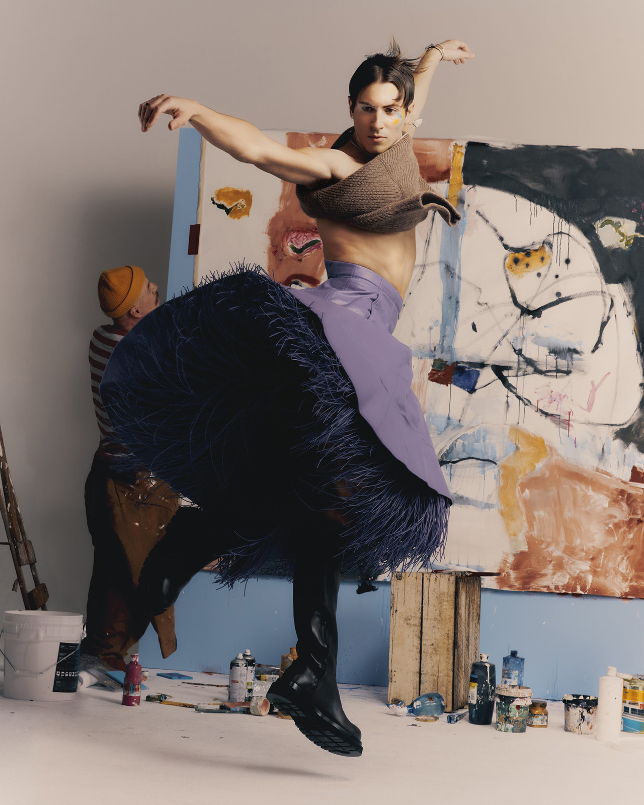 Man in avant-garde fashion leaps in an art studio with a striped-shirt artist, paint cans, and an abstract painting in the background.