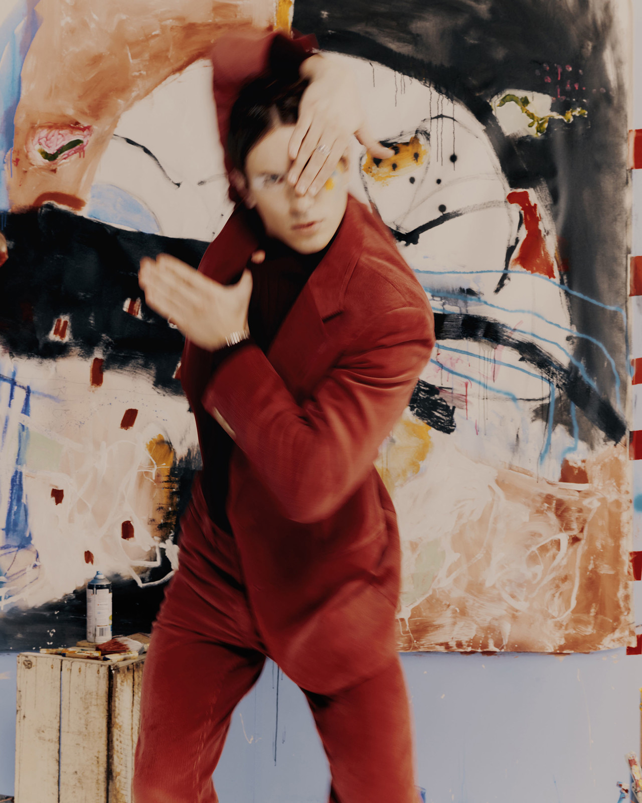 Young man in a red suit poses in front of a large, colourful abstract painting in an artist's studio, obscuring his face with his hands.