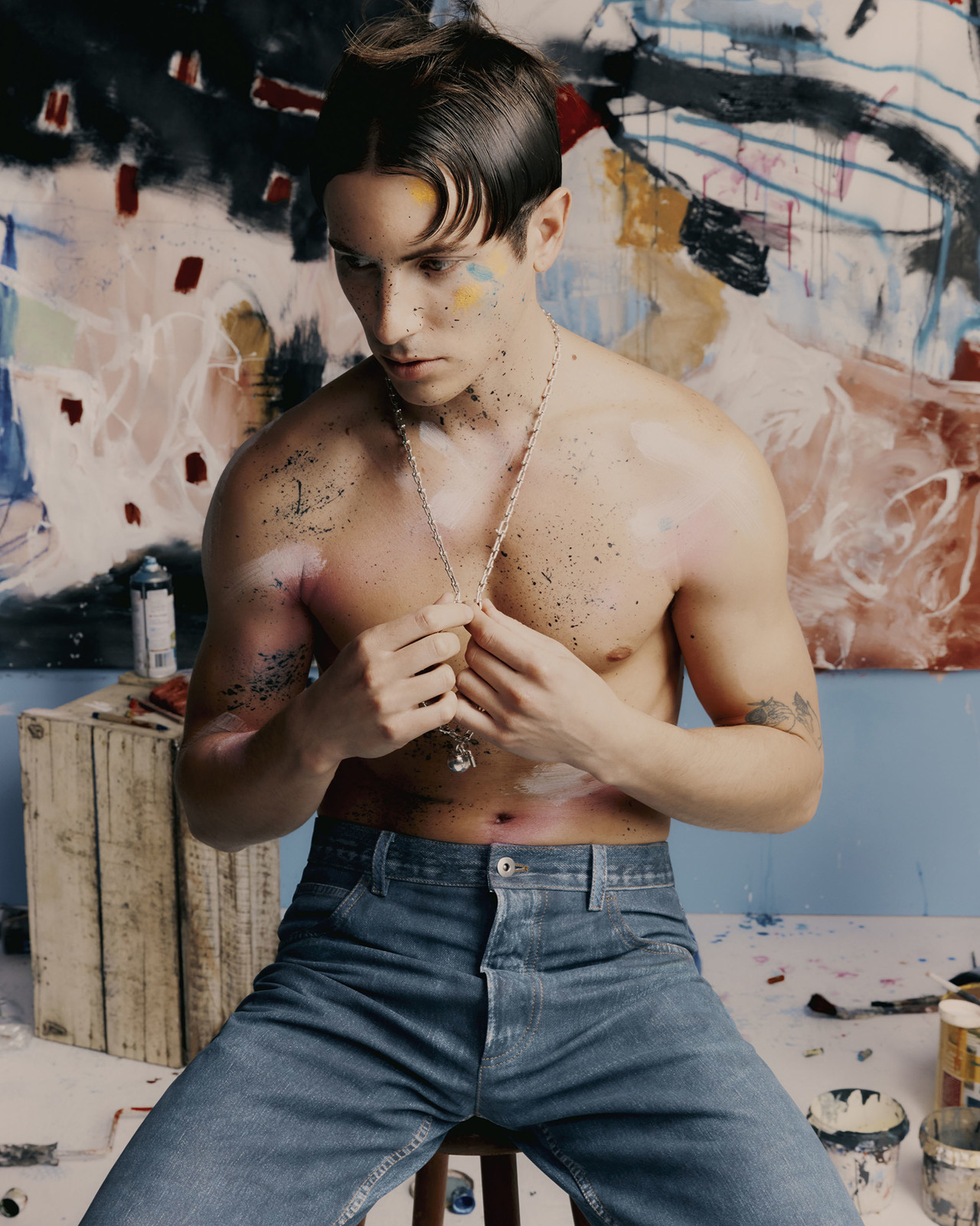 Shirtless man with paint on his face and body sits on a stool in front of a paint-splattered backdrop, wearing blue jeans and a silver necklace.