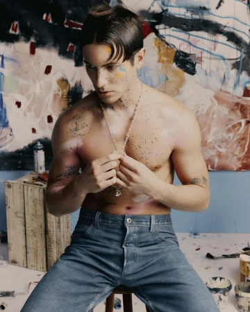 Shirtless man with paint on his face and body sits on a stool in front of a paint-splattered backdrop, wearing blue jeans and a silver necklace.
