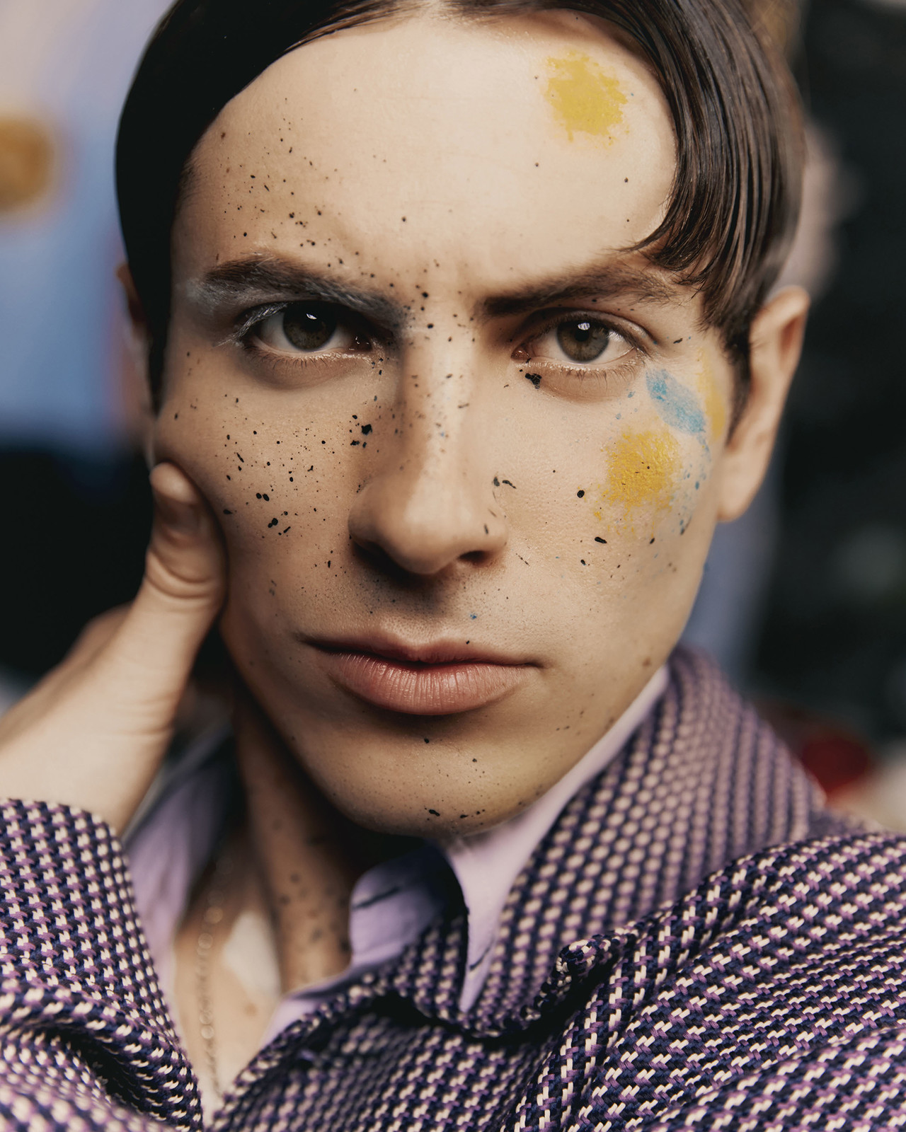 Close-up portrait of a young adult male with dark hair and light skin, posing with hand on face, wearing a purple textured blazer.