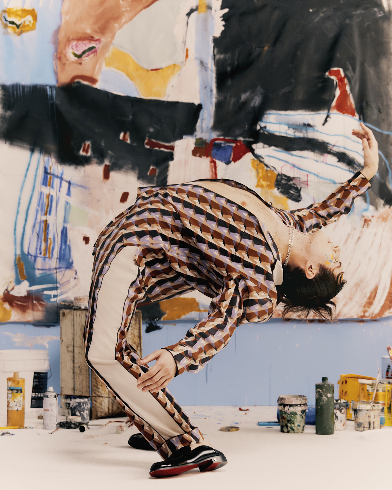 Adult in patterned shirt arches back in an art studio filled with paint, standing before a colorful abstract painting in a fashion editorial.