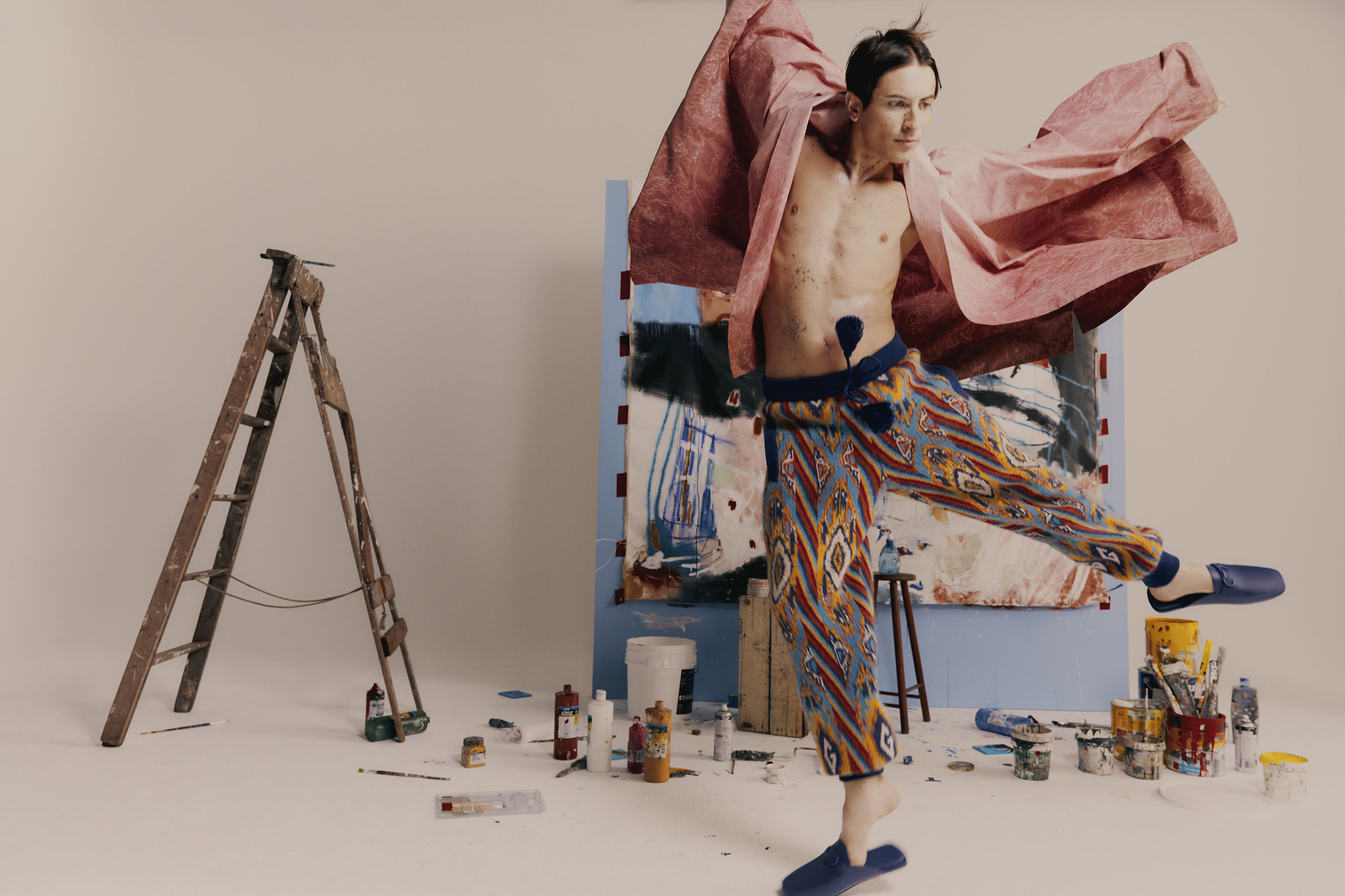 Adult male in patterned pants jumps in a studio filled with paint cans, a ladder, paint splatters, and an abstract painting.