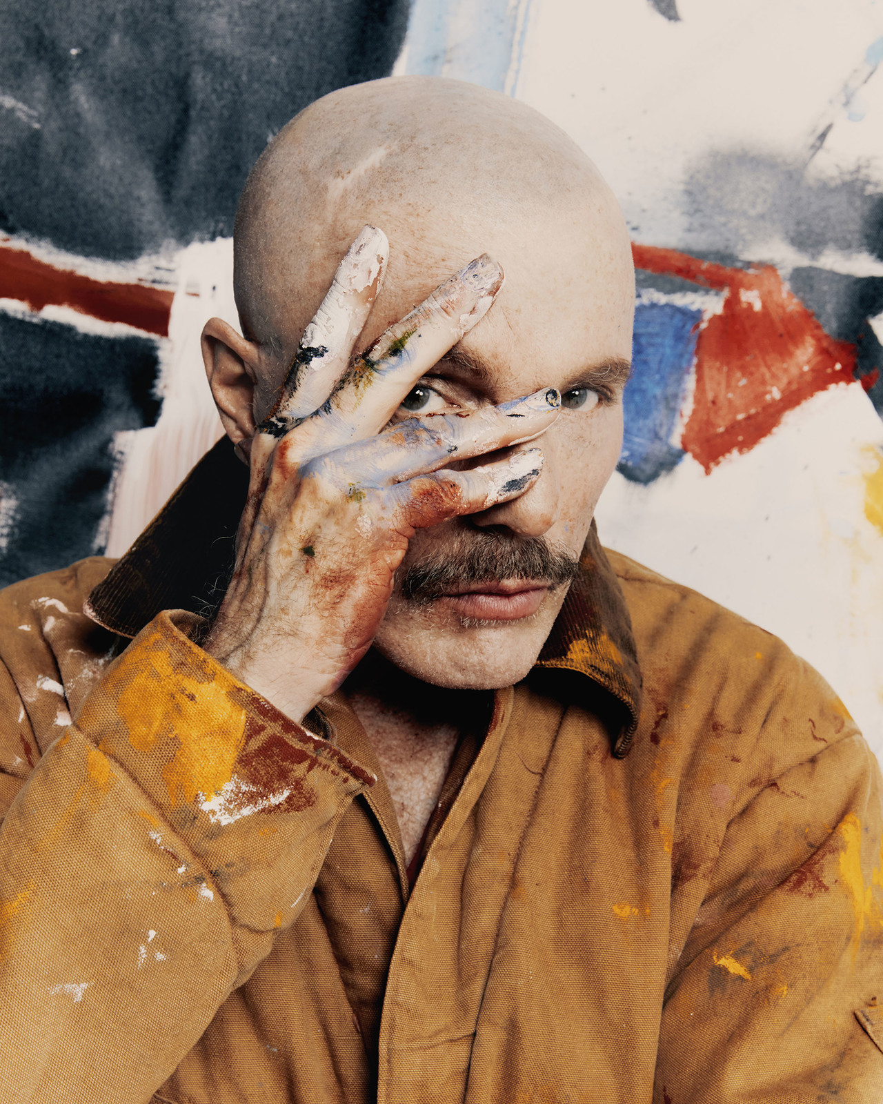Bald man with mustache covers one eye with paint-covered hand against a blurry backdrop with brown jacket and intense gaze.