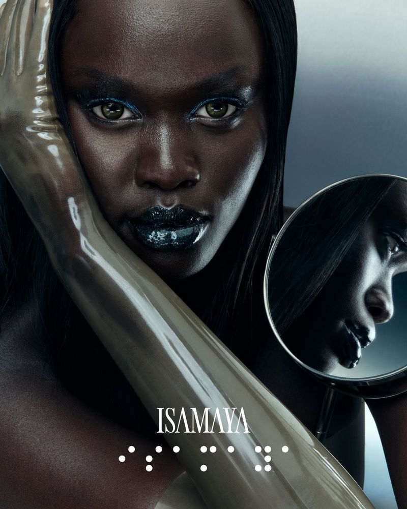 A dark-skinned woman with glossy dark lipstick, latex-like sleeves, and a circular mirror reflecting her face, against a grey background.