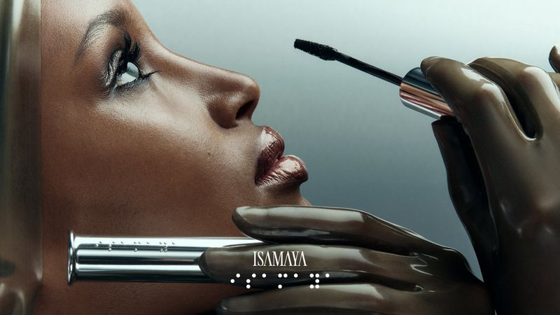 Close-up of a woman's face with dark skin and glossy lips getting mascara applied by hands in brown gloves, cosmetics ad for ISAMAYA.