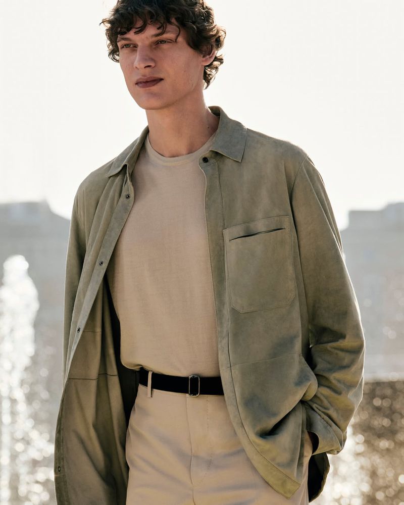 Outdoor fashion portrait of a young adult male wearing a light green button-up shirt, beige t-shirt, and pants, standing near a fountain.
