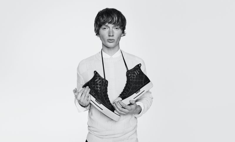Monochrome studio shot of a young adult man holding a pair of black high-top sneakers against a plain white backdrop.