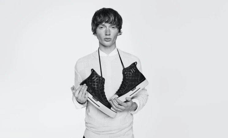 Monochrome studio shot of a young adult man holding a pair of black high-top sneakers against a plain white backdrop.
