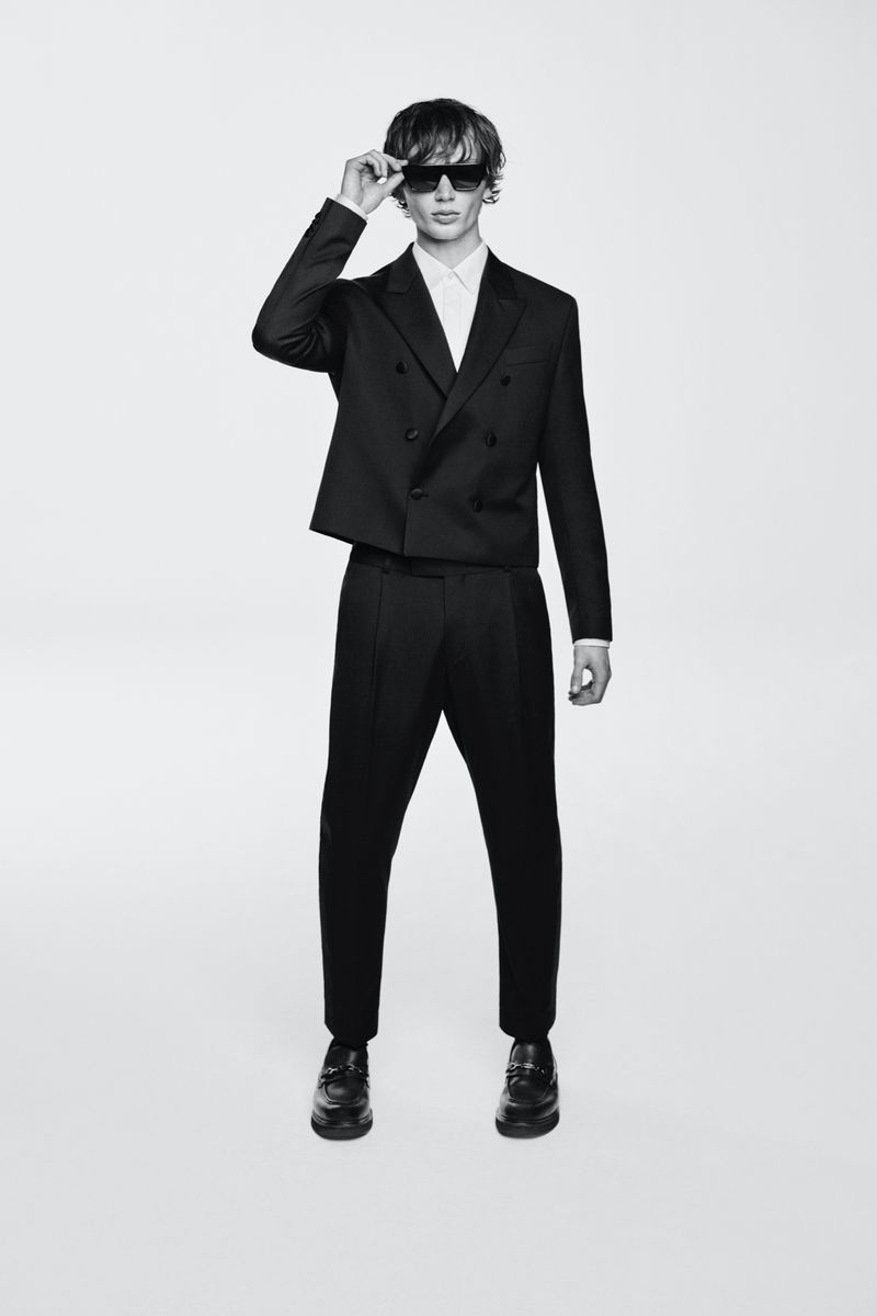 Model wearing a black double-breasted suit and sunglasses adjusting glasses against a seamless white background.