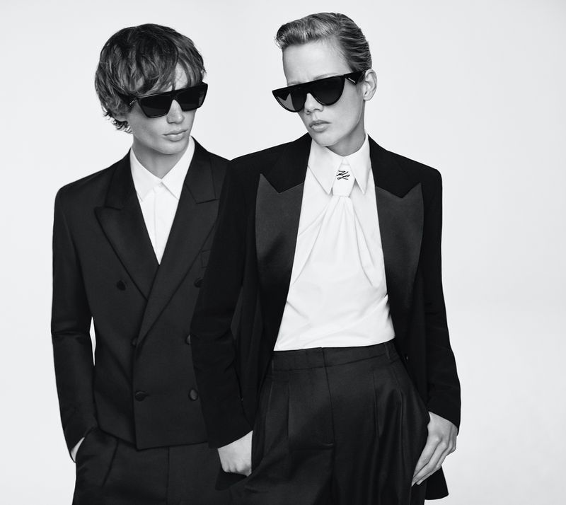 Two fashion models pose in black tuxedos and sunglasses against a bright background, conveying a sleek and stylish aesthetic.