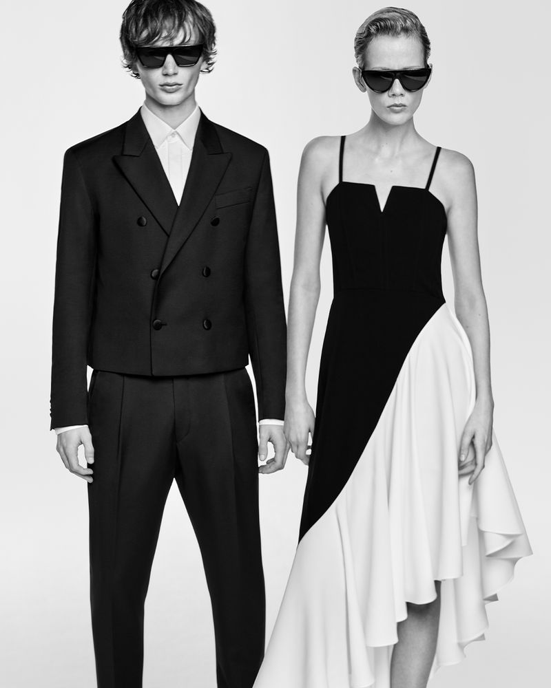 A stylish black-and-white fashion shot featuring an adult male model in a suit and an adult female model in a dress, both wearing sunglasses.