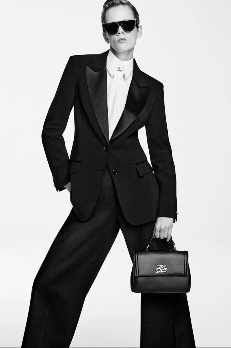 A stylish woman in a black pantsuit and sunglasses holds a black handbag against a neutral backdrop in a studio fashion shot.