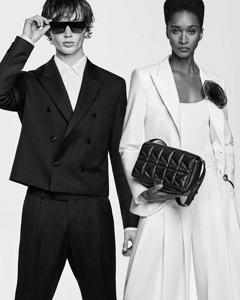 Two fashion models, one male and one female, pose in a studio wearing stylish black and white suits with a handbag in hand.