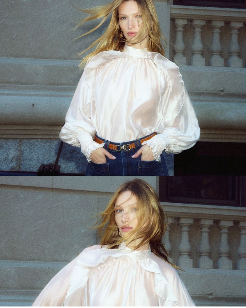 An adult woman poses on concrete stairs in a translucent blouse and jeans. She is wearing a brown belt and gold jewelry. Her blonde hair is blowing.