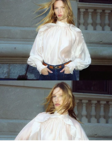 An adult woman poses on concrete stairs in a translucent blouse and jeans. She is wearing a brown belt and gold jewelry. Her blonde hair is blowing.