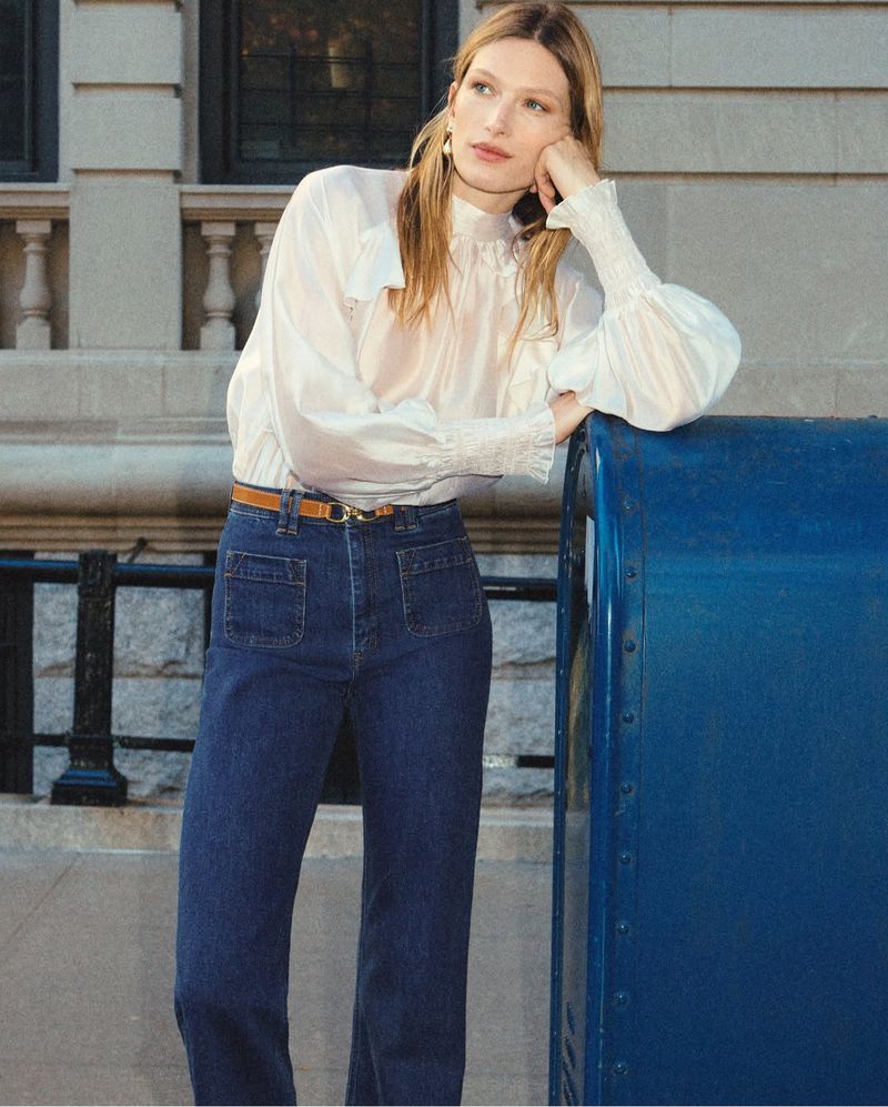 Woman with long blond hair stands by blue mailbox wearing a white blouse with long sleeves and blue jeans in an outdoor urban setting.
