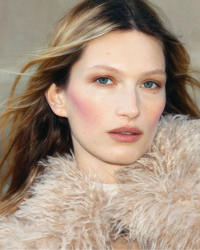 Close-up portrait of a fair-skinned young adult female with blonde hair and blue eyes wearing a cream-colored, furry jacket against a plain background.