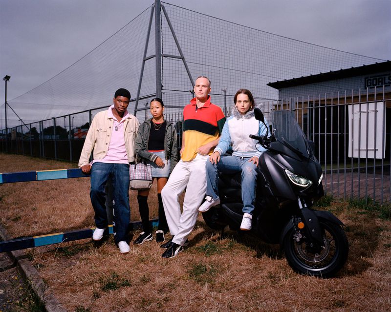 Four young adults posing near a motorcycle and soccer field, wearing casual outfits, with a muted color palette and an overcast sky.