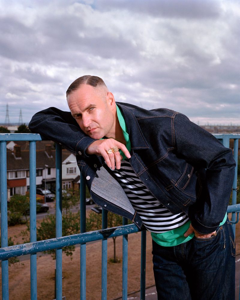 Adult male leaning on a rail outdoors wearing a dark denim jacket, striped shirt, and jeans, with houses and cloudy sky in background.