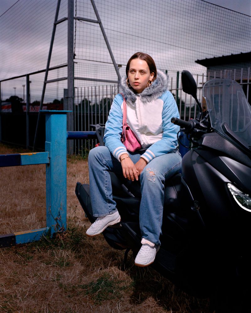 Young woman sitting on a black motor scooter, wearing a blue and white jacket, light blue jeans, white sneakers, in an outdoor setting.