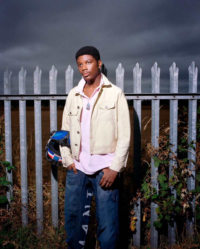 Young African-American man standing outdoors in jacket and jeans holding helmet, posing in front of fence with field and cloudy sky.
