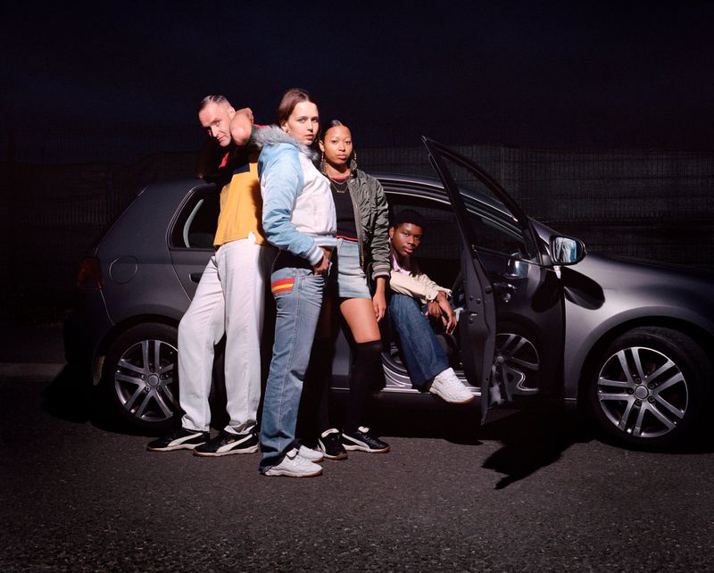 Group of four young adult friends pose in front of a silver hatchback car at night, illuminated by bright light.