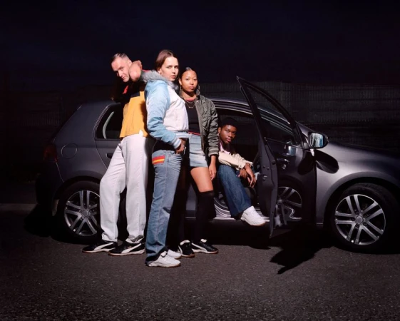 Group of four young adult friends pose in front of a silver hatchback car at night, illuminated by bright light.