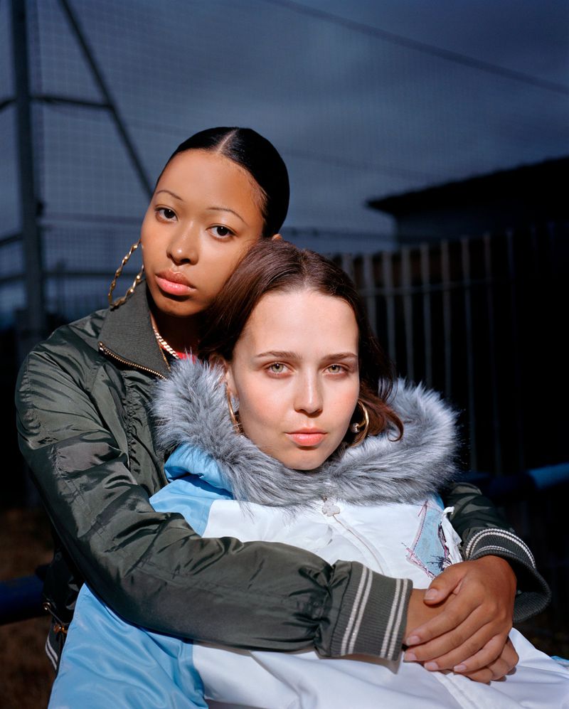 Two young women pose outdoors at dusk, one with tan skin and an olive-green jacket, the other in a blue and white hooded parka.