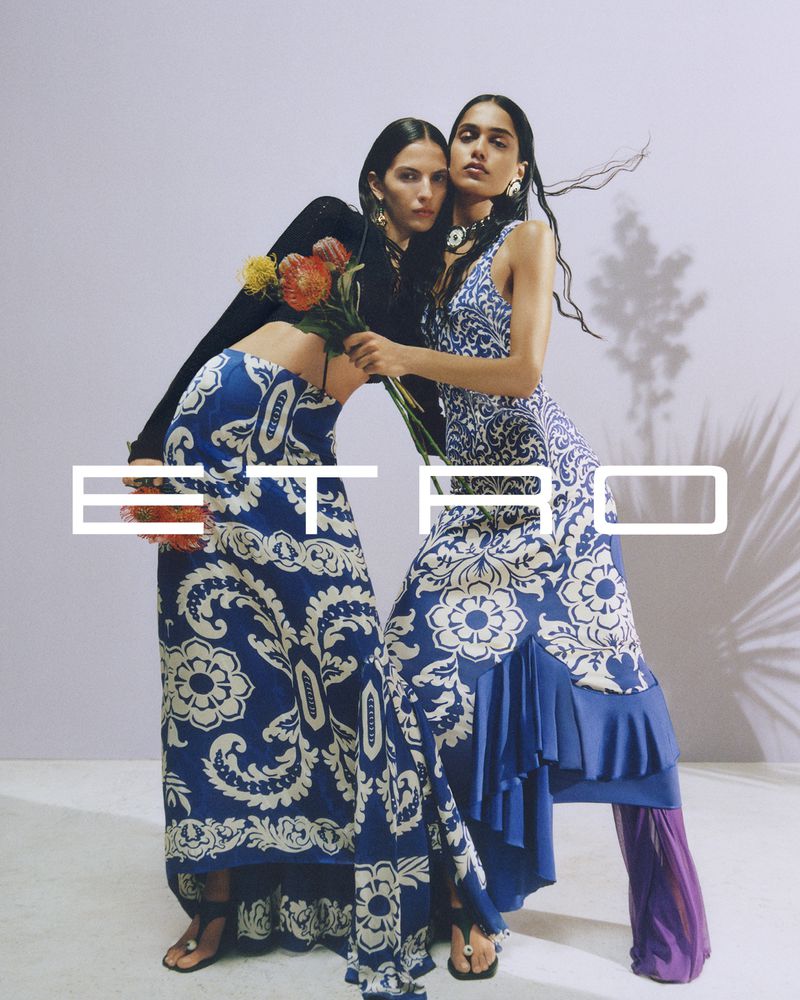 Two women pose in patterned Etro maxi dresses holding flowers, illuminated by soft light with subtle shadows. Fashion shoot.