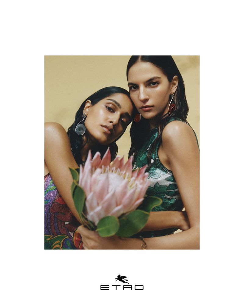Two adult females pose indoors with a large pink protea flower. One woman wears a sequin dress. Fashion, beauty and elegant jewelry visible.