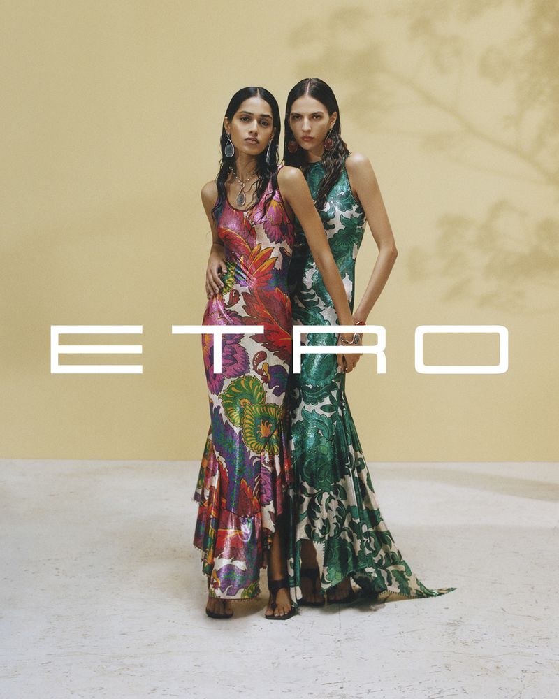 Two fashion models pose closely in floral-print dresses, with a blurred tree shadow and Etro logo overlay, against a soft yellow backdrop.