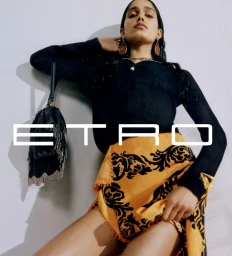 Model in black top and patterned yellow skirt holding a fringed handbag against a white studio wall