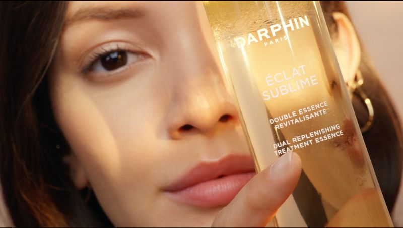 Close-up of an adult woman holding a Darphin ÉCLAT SUBLIME Double Essence Revitalisante bottle; beauty and skincare advertisement.