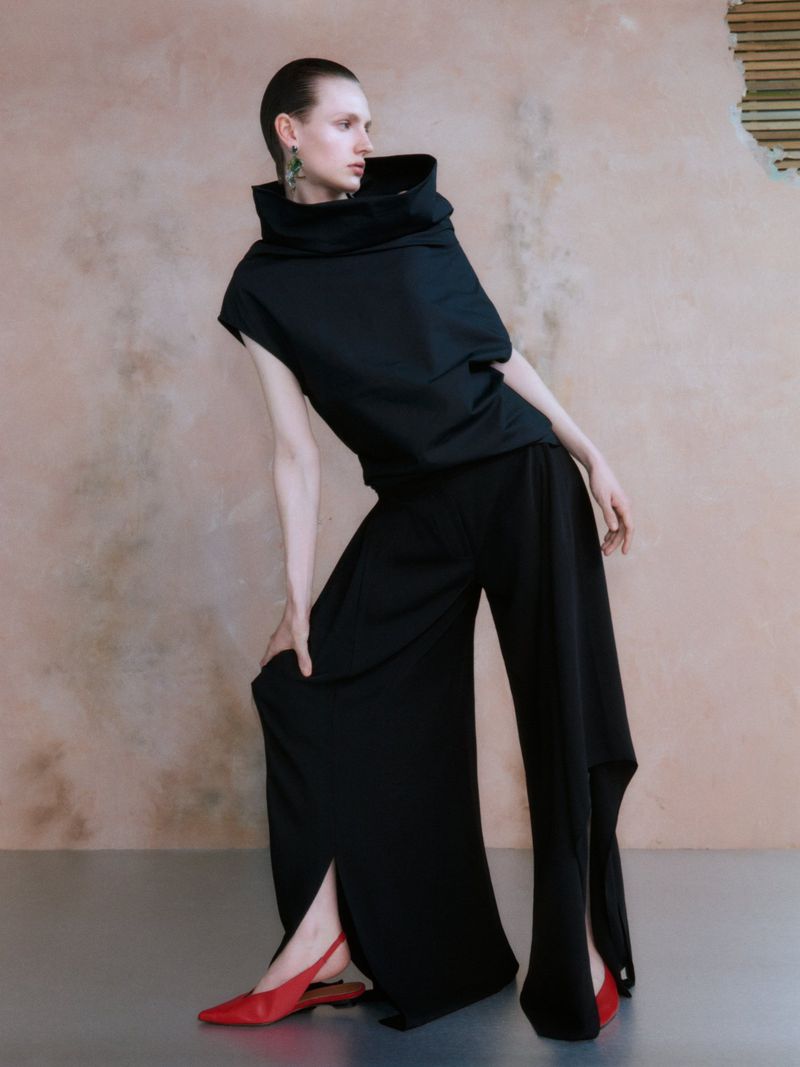 Woman in flowing black top and pants poses in photo studio with artistic flair, wearing red shoes and green earrings against a textured wall.