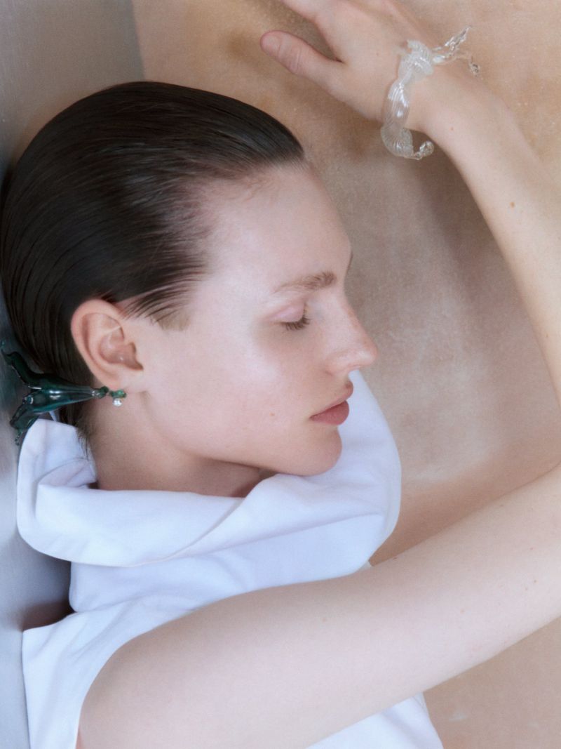 Profile portrait of a woman with slicked-back dark hair and a white collar against a neutral textured background for Qasimi SS25.