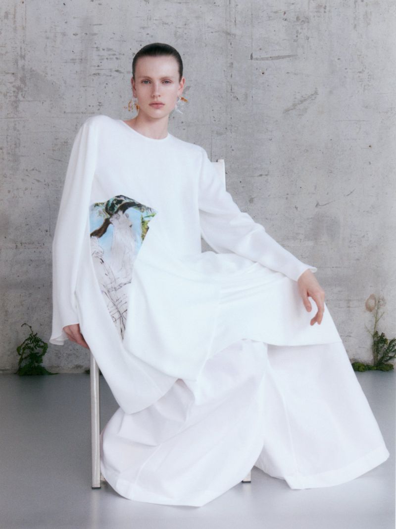 A person with short brown hair in a white dress sits in a chair in front of a light grey concrete wall with subtle greenery.
