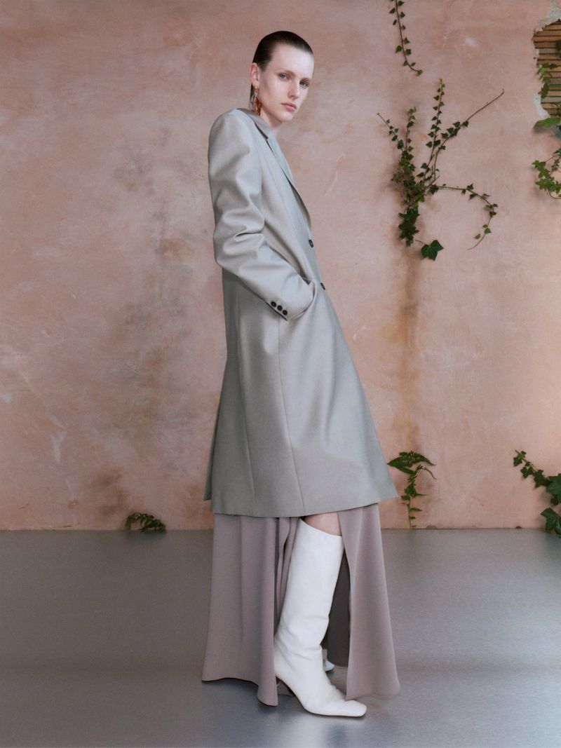 Elegant fashion shot of a woman in a long grey coat, dress and white boots, posing in front of a neutral backdrop with vines.
