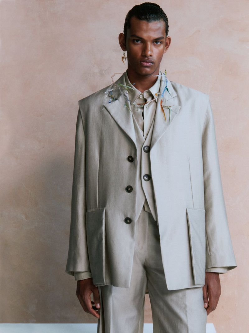 Dark-skinned male fashion model in a light beige three-piece suit poses indoors against a light-textured backdrop with soft lighting.