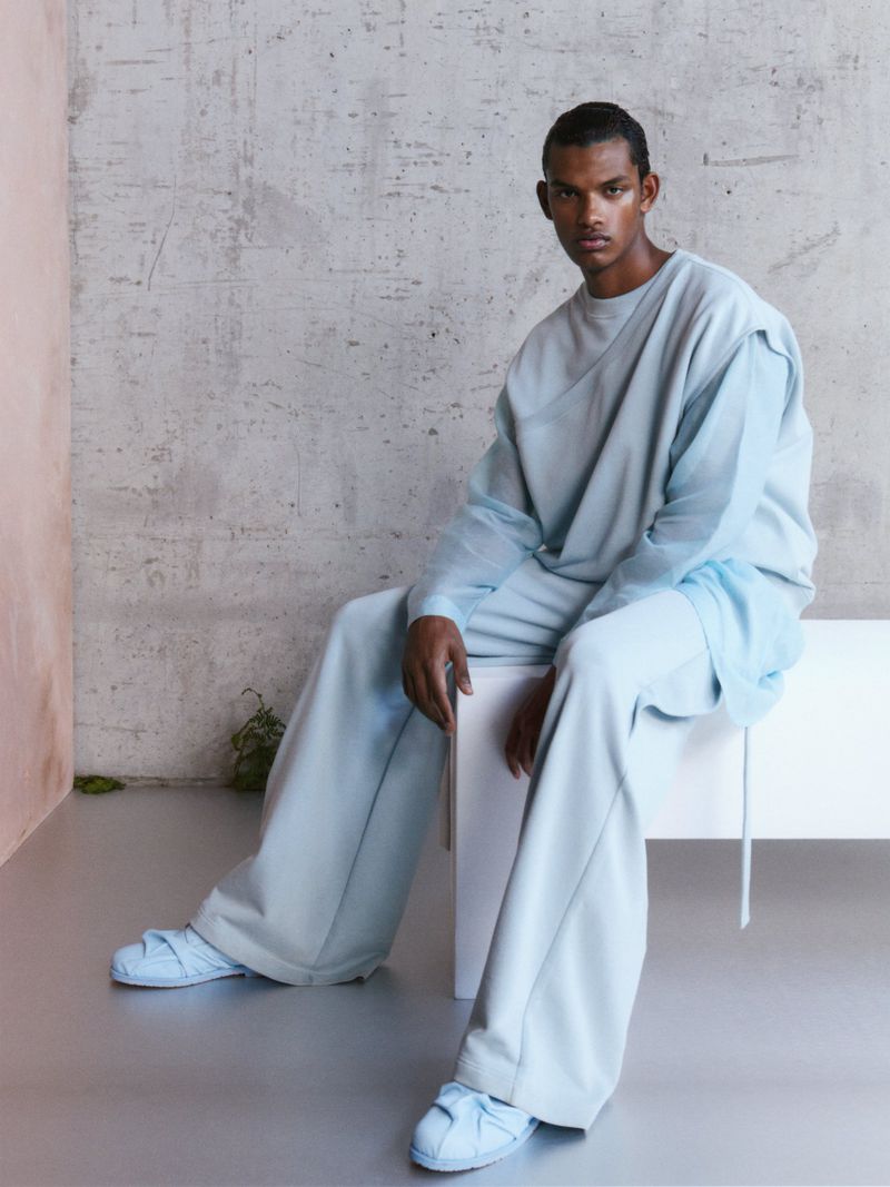 A dark-skinned male model is seated on a white bench wearing an ice-blue oversized lounge set with matching slide sandals in a studio setting.