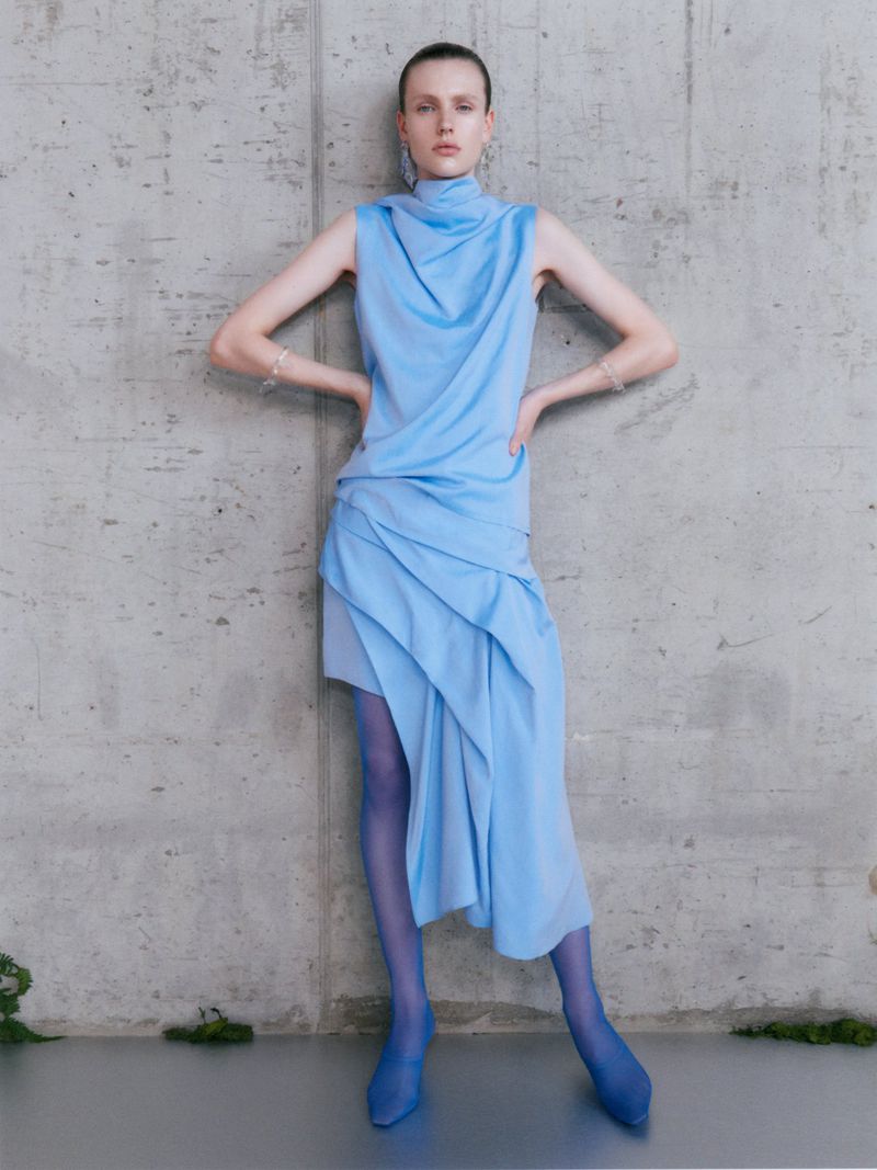 A poised female model showcases a light blue draped dress with matching stockings and heels against a textured grey wall in a fashion editorial.