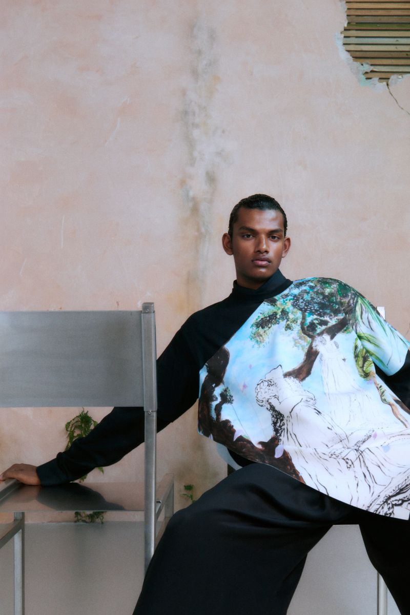 Adult male in black top and pants poses while seated on a metal chair in front of a pale pink wall in a fashion setting.