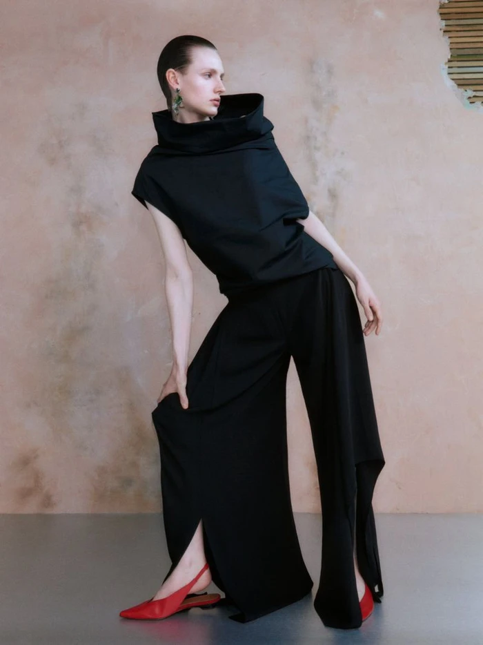 Model in a black cowl-neck top and wide-leg trousers posing against a textured wall with red shoes.