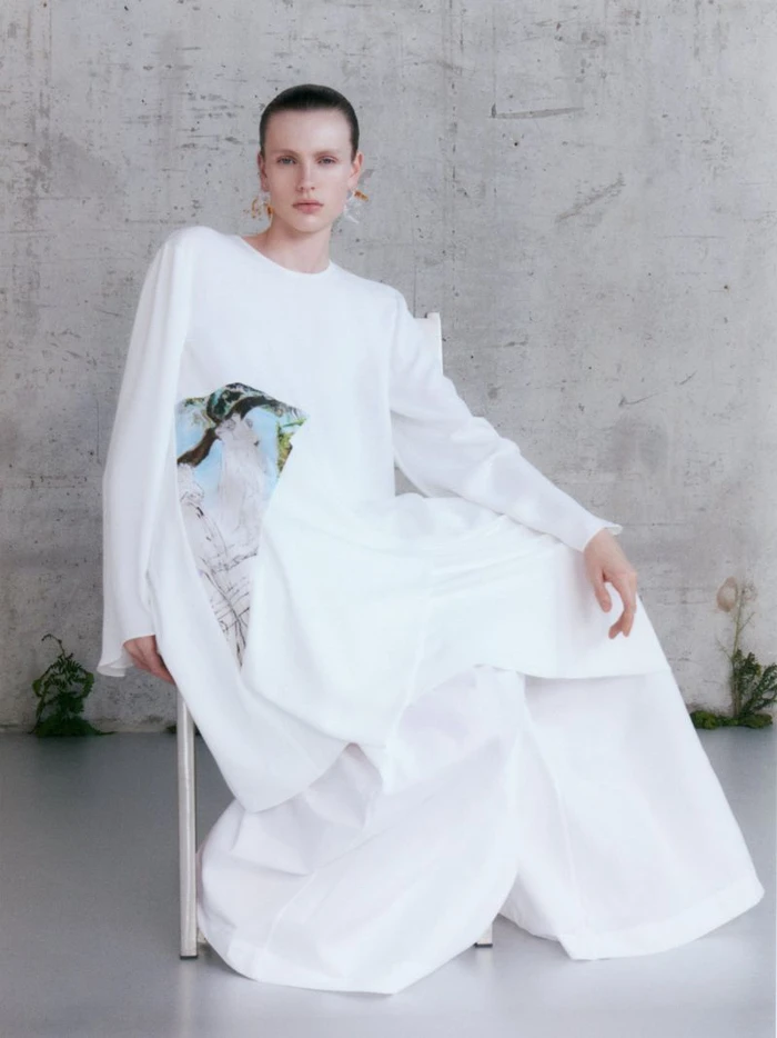 Model wearing a white dress with a graphic print seated on a chair against a textured concrete wall.