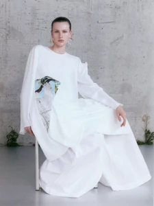 Model wearing a white dress with a graphic print seated on a chair against a textured concrete wall.