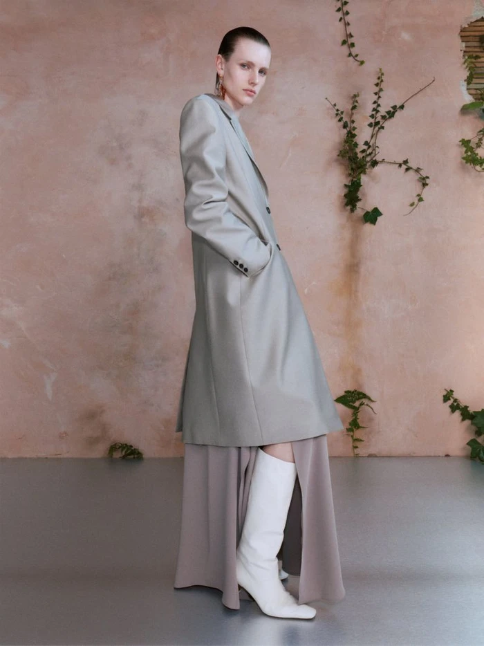 Model wearing a grey coat and white boots against a textured pink wall with climbing vines for Qasimi Spring/Summer 2025.