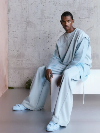 Man wearing a light blue crewneck sweater and matching trousers seated against a textured concrete wall.