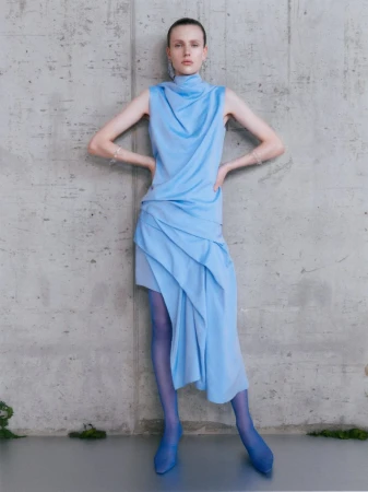 Model in a draped light blue sleeveless top and skirt standing against a concrete wall for Qasimi