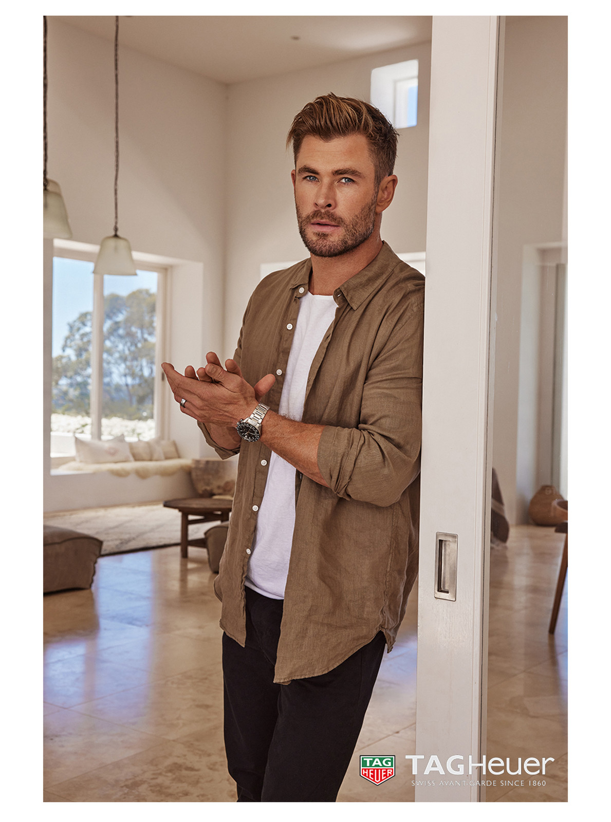 Adult male poses indoors wearing a button-down shirt, t-shirt, and pants, accessorized with a wristwatch while standing by a white door.
