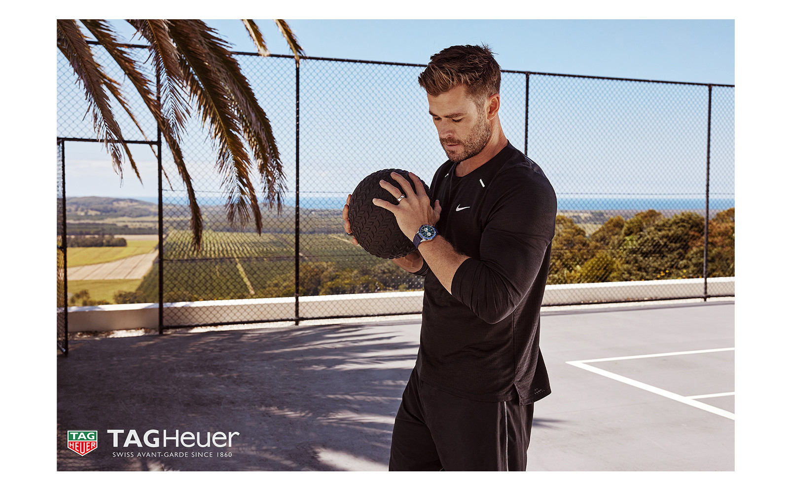 Athletic man in black activewear poses outdoors, holding a medicine ball; a luxury watch is visible on his wrist against a natural backdrop.